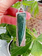Green stone pendant with silver frame held by a hand against green leaves