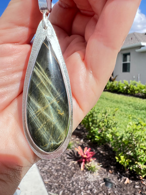A hand holding a teardrop-shaped pendant featuring a polished green stone with gold veining, set in a textured silver frame, with a sunny backyard garden in the background.